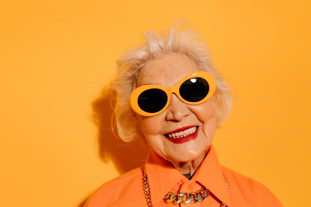 Portrait of good looking middle aged woman with wrinkled face and creative sunglasses dressed in stylish clothes looking directly at the camera with calm expression isolated on yellow studio wall