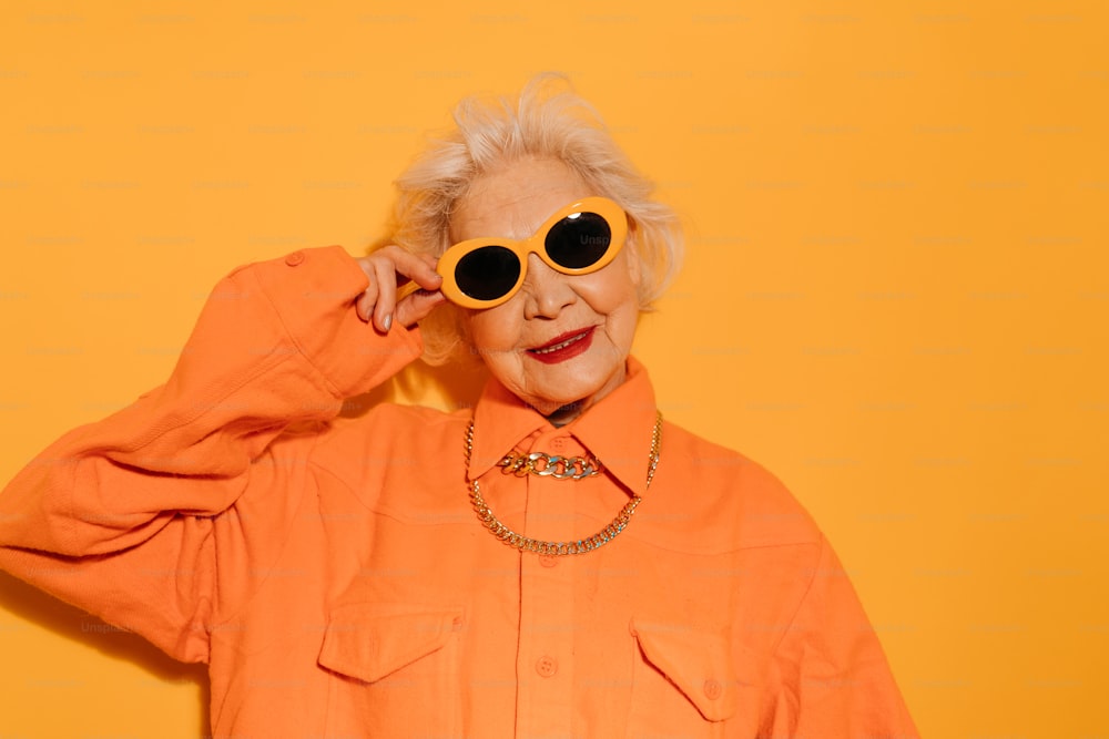 Stylish. Horizontal view of the senior woman with grey hair wearing sunglasses dressed in fashionable clothes looking at the camera âwhile posing at the studio with yellow wall. Stock photo