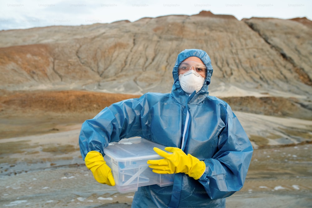 Jeune chercheuse en combinaison de protection, respirateur, gants en caoutchouc et lunettes tenant un récipient en plastique avec des échantillons d’eau et de sol