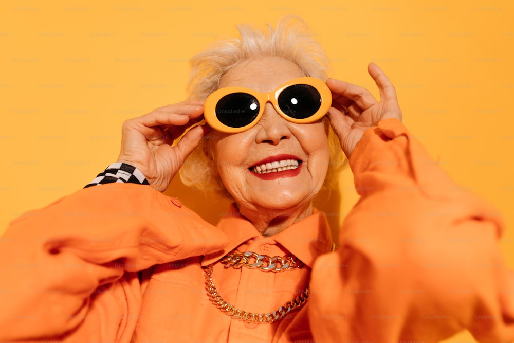Portrait view of the senior woman with grey hair dressed in fashionable clothes smiling to the camera âand holding hands at her sunglasses while posing at the studio with yellow wall. Stock photo