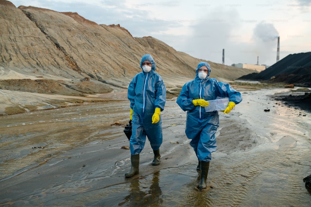 Two young contemporary female scientists in blue protective coveralls and rubber boots moving down polluted river surrounded by hills