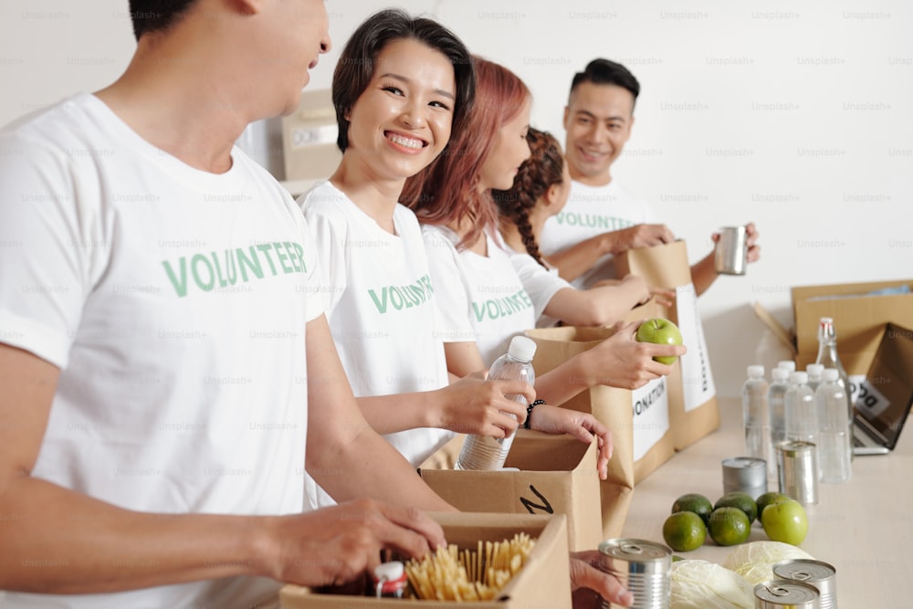 Group of smiling young students volunteering at charitable foundation office and packing food and waterr in big paper packages for people in need