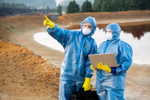 Junge Forscherin zeigt nach vorne, während sie einem Kollegen mit Laptop zeigt, wo er Proben von giftigem Boden und Wasser entnehmen soll