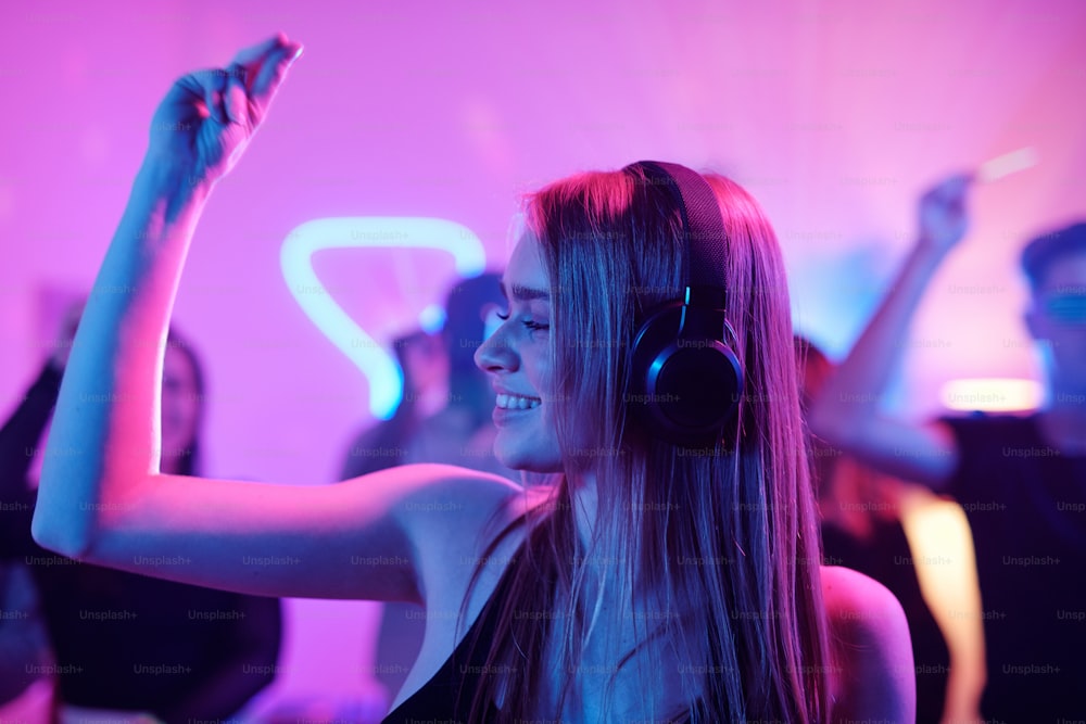 Jovem mulher alegre com longos cabelos loiros estalando os dedos enquanto desfruta de música em fones de ouvido na frente da multidão dançante na festa
