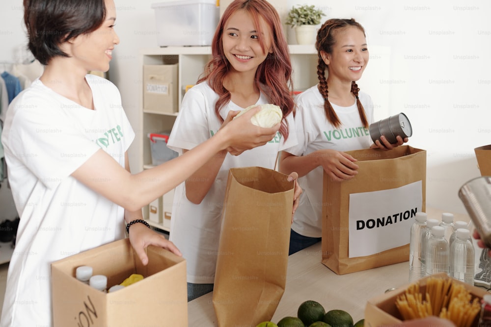 Lächelnde junge asiatische Frauen, die Konserven, Obst, Gemüse und Wasserwasser in Papierverpackungen verpacken, um in einem Wohltätigkeitszentrum zu spenden