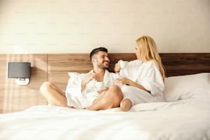 Talking in bed, a morning gossip party with your loved one over a cup of hot beverage, coffee or tea. Adult man and woman in the robes are comfortably placed in a bed and they laugh at each other