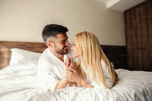 Waking up with a smile, mornings with a loved one. Sunny mornings in a room of a modern hotel, close couple in white bathrobes share a moment to remember. He holds her hands and looks her in the eye