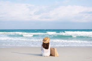Imagen de vista trasera de una mujer con sombrero y bolso sentada en la playa con fondo azul cielo