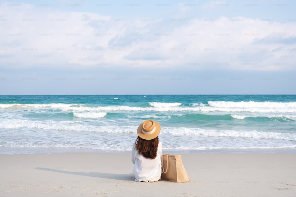 Rear view image of a woman with hat and bag sitting on the beach with blue sky background