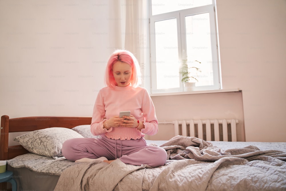 Enjoying with gadgets. Full length view of the woman with pink hair sitting at the bed and typing something at her smartphone at the morning. Stock photo