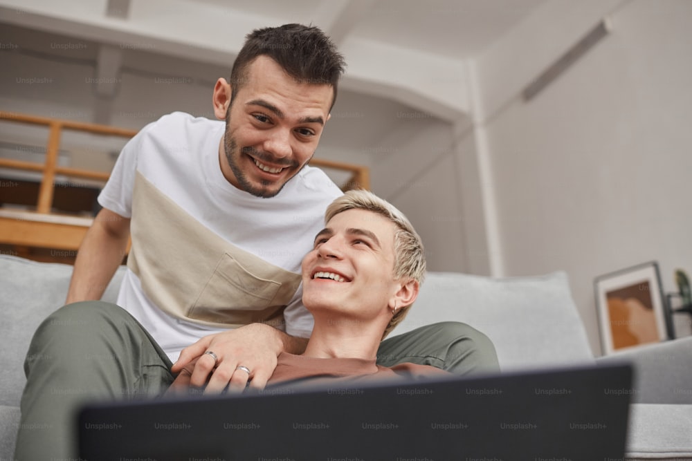 Vista a bassa angolazione alla coppia gay maschile che sorride felicemente mentre usa il laptop insieme a casa, copia lo spazio