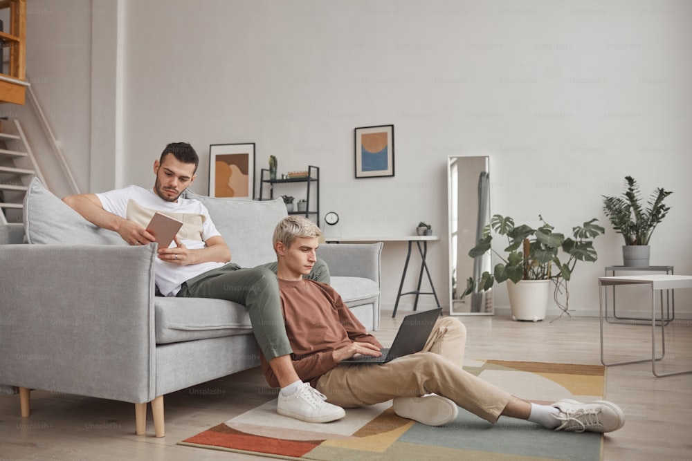 Ritratto a figura intera di coppia gay contemporanea che utilizza i computer mentre si rilassa sul divano insieme in interni domestici minimali, spazio di copia