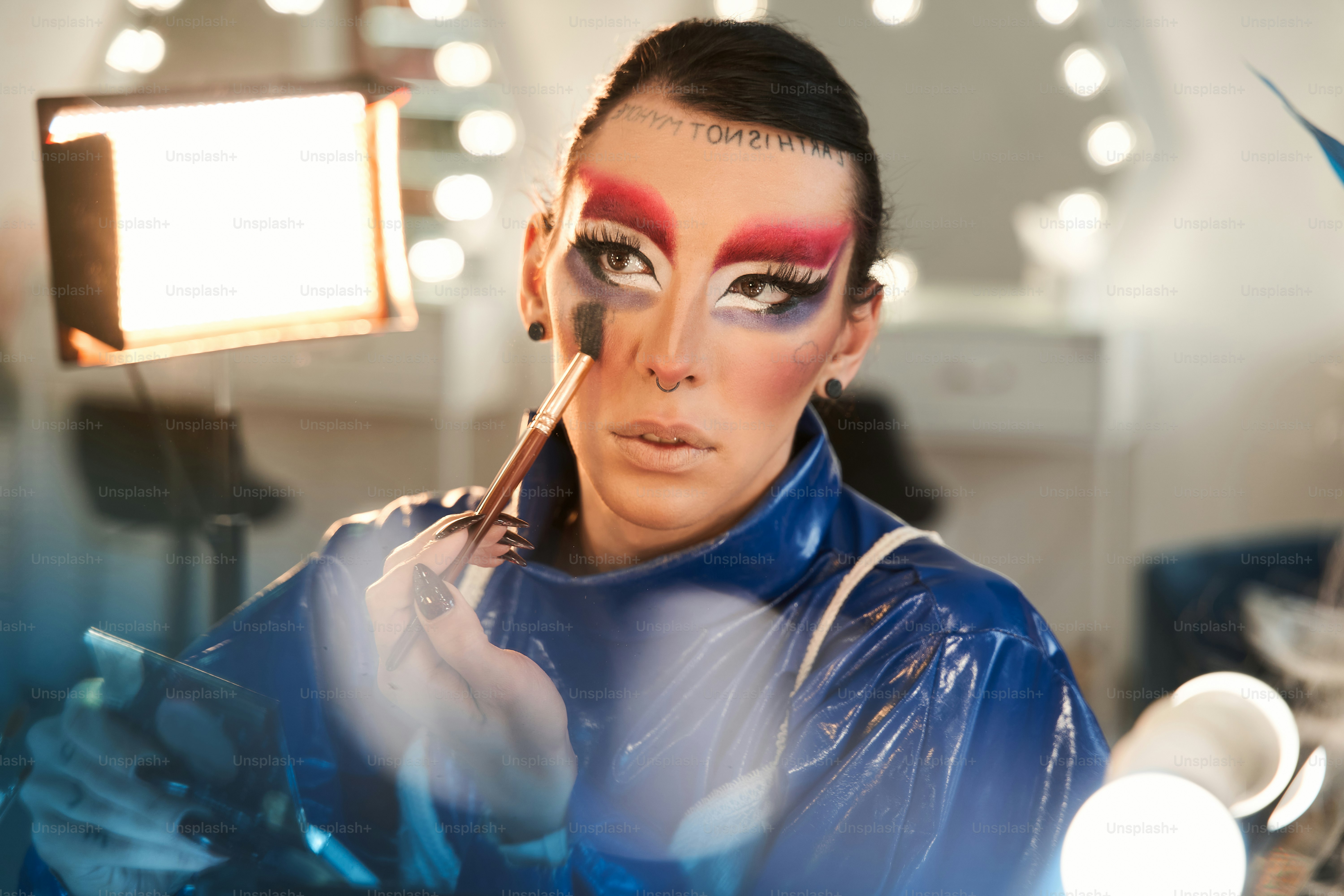 Portrait view of the drag queen with false eyelashes looking at the mirror and applying paints at his face while sitting at the dressing table. Drag queen and transgender person concept