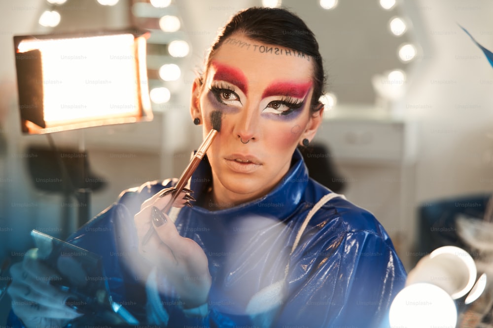 Portrait view of the drag queen with false eyelashes looking at the mirror and applying paints at his face while sitting at the dressing table. Drag queen and transgender person concept