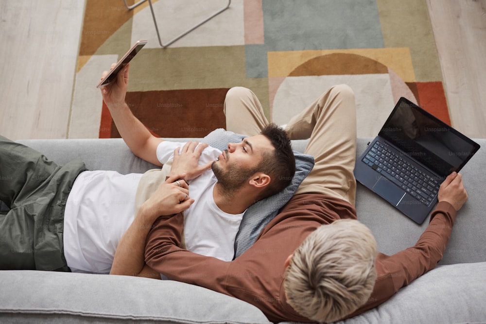 Top view at contemporary gay couple using computers while relaxing on couch together in minimal home interior, copy space