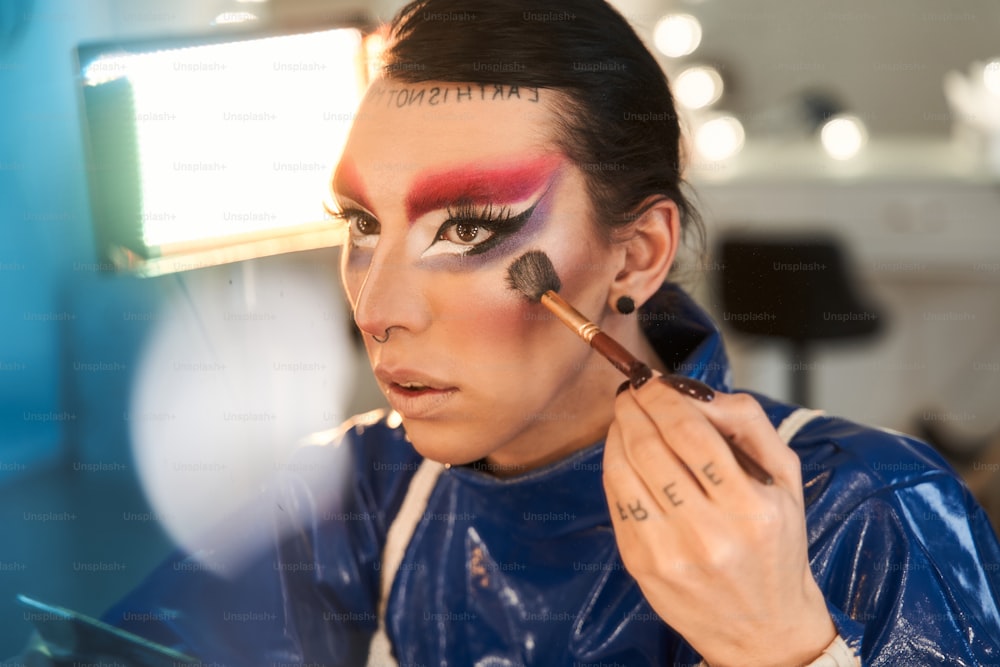 Vista retrato da drag queen com maquiagem brilhante aplicando marca-texto em seu rosto enquanto está sentada na penteadeira. Conceito de drag queen e pessoa transgênero