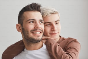 Retrato sincero de um casal gay despreocupado abraçando-se dentro de casa e olhando para longe feliz enquanto posa contra branco, espaço de cópia