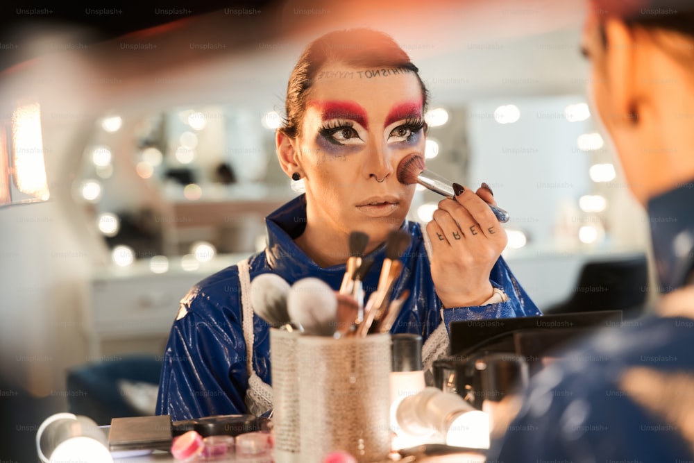 Vista ritratto della drag queen che guarda lo specchio e applica arrossimenti sul suo viso mentre è seduta al tavolo da toeletta. e preparando la sua reincarnazione. âConcetto di drag queen e persona transgender