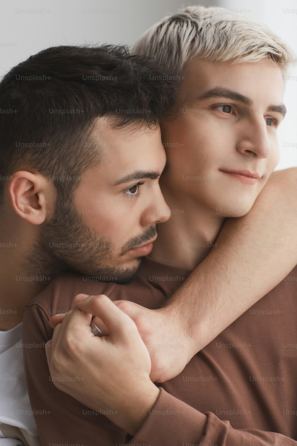 Vertical close up portrait of contemporary gay couple embracing tenderly and holding hands while looking away