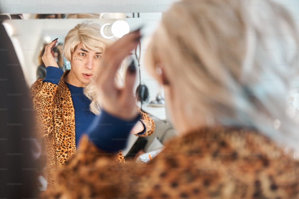 Rückansicht der Dragqueen, die vor dem Spiegel sitzt und sich eine Perücke auf den Kopf setzt, während sie seinen neuen Look vorbereitet. Dragqueen und Transgender-Person-Konzept. Stockfoto
