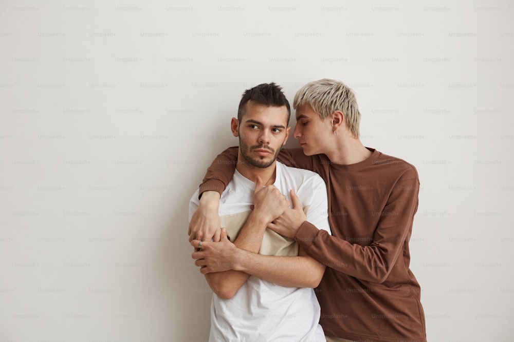 Ritratto minimale della vita in su di una giovane coppia gay che si abbraccia mentre si trova contro il muro bianco all'interno, spazio di copia