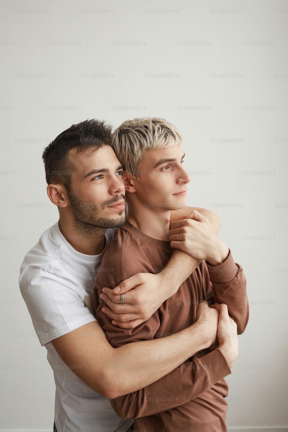 Vertical waist up portrait of loving gay couple embracing at home and looking away while standing against white wall, copy space