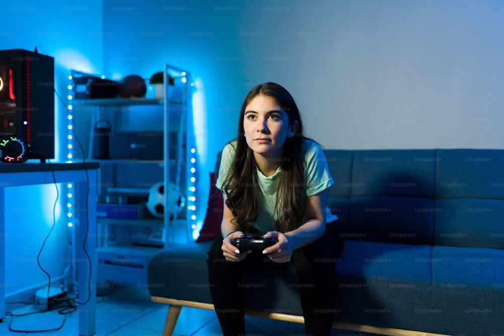 Gut aussehende junge Frau, die mit LED-Leuchten auf der Couch in ihrem Schlafzimmer sitzt, während sie eine Fernbedienung hält und ein Videospiel spielt