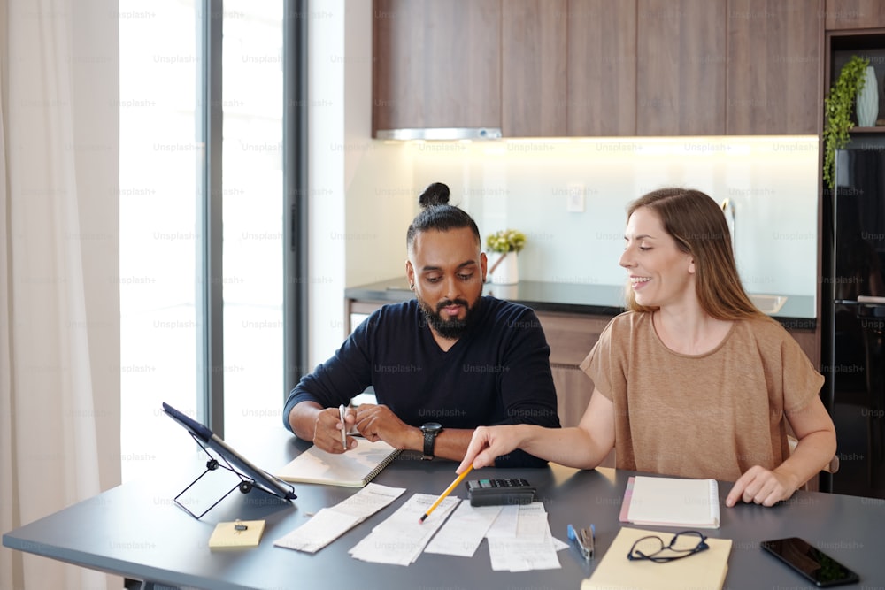 Smiling young woman pointing at bills on table and explaining boyfrend how to manage budget