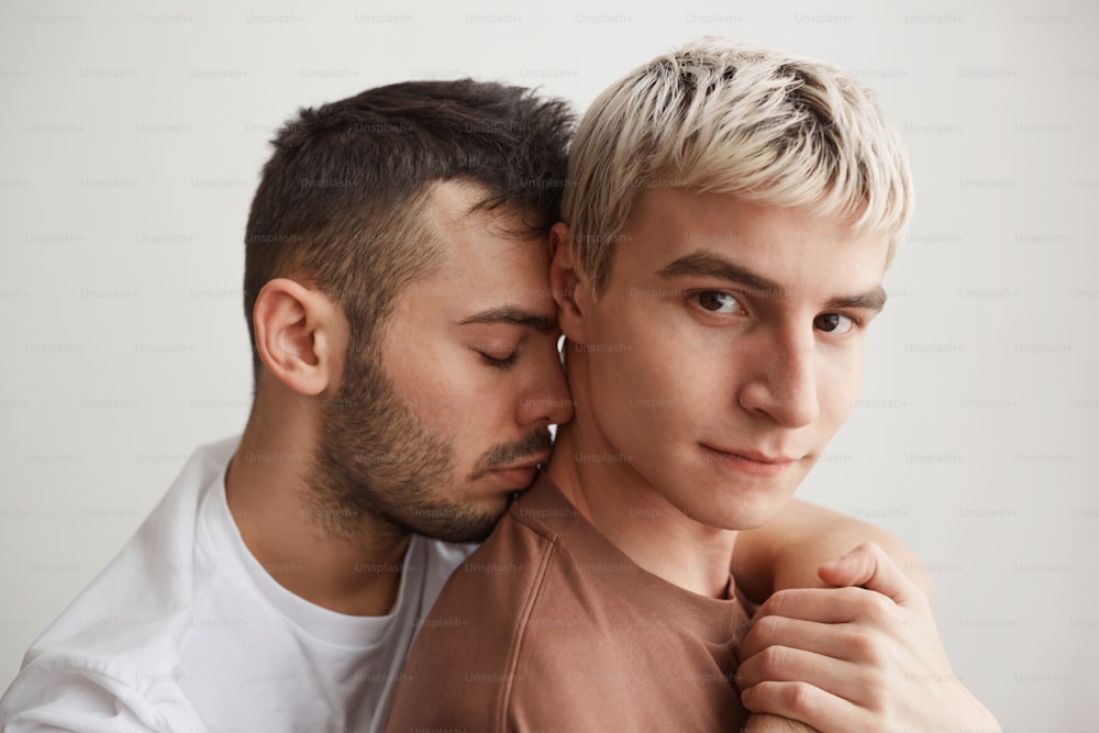 Ritratto ravvicinato di coppia gay amorevole che abbraccia e guarda la telecamera mentre si trova contro il bianco a casa
