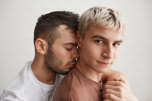 Close up portrait of loving gay couple embracing and looking at camera while standing against white at home