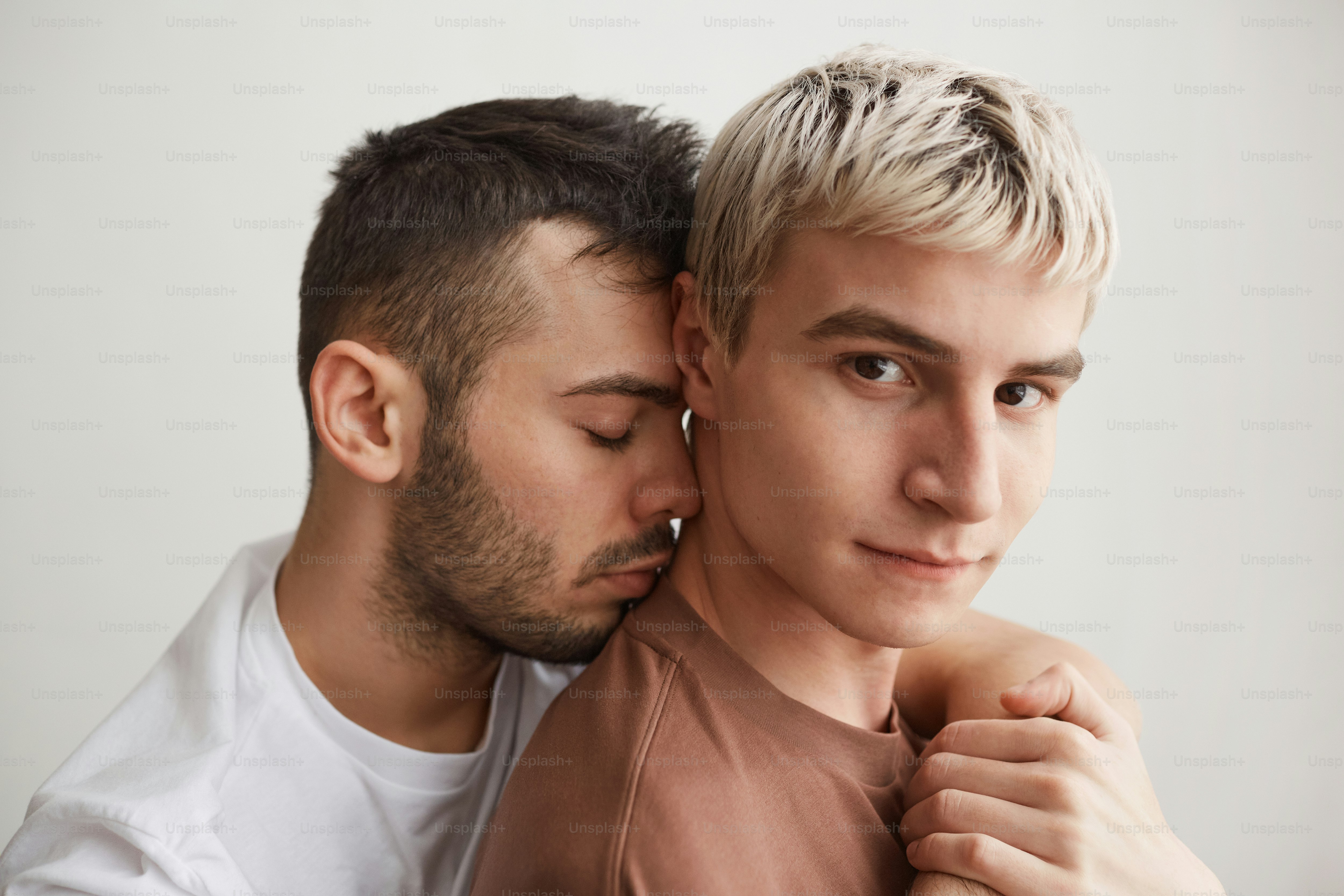 Close up portrait of loving gay couple embracing and looking at camera while standing against white at home