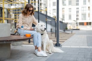 Cheerful woman sitting on bench with big white dog in the courtyard of the residence while using phone. Animal lover, pet friendly owner.