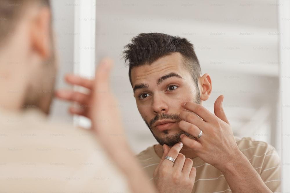 Close up of young man applying face cream while looking in mirror during morning skincare routine, copy space