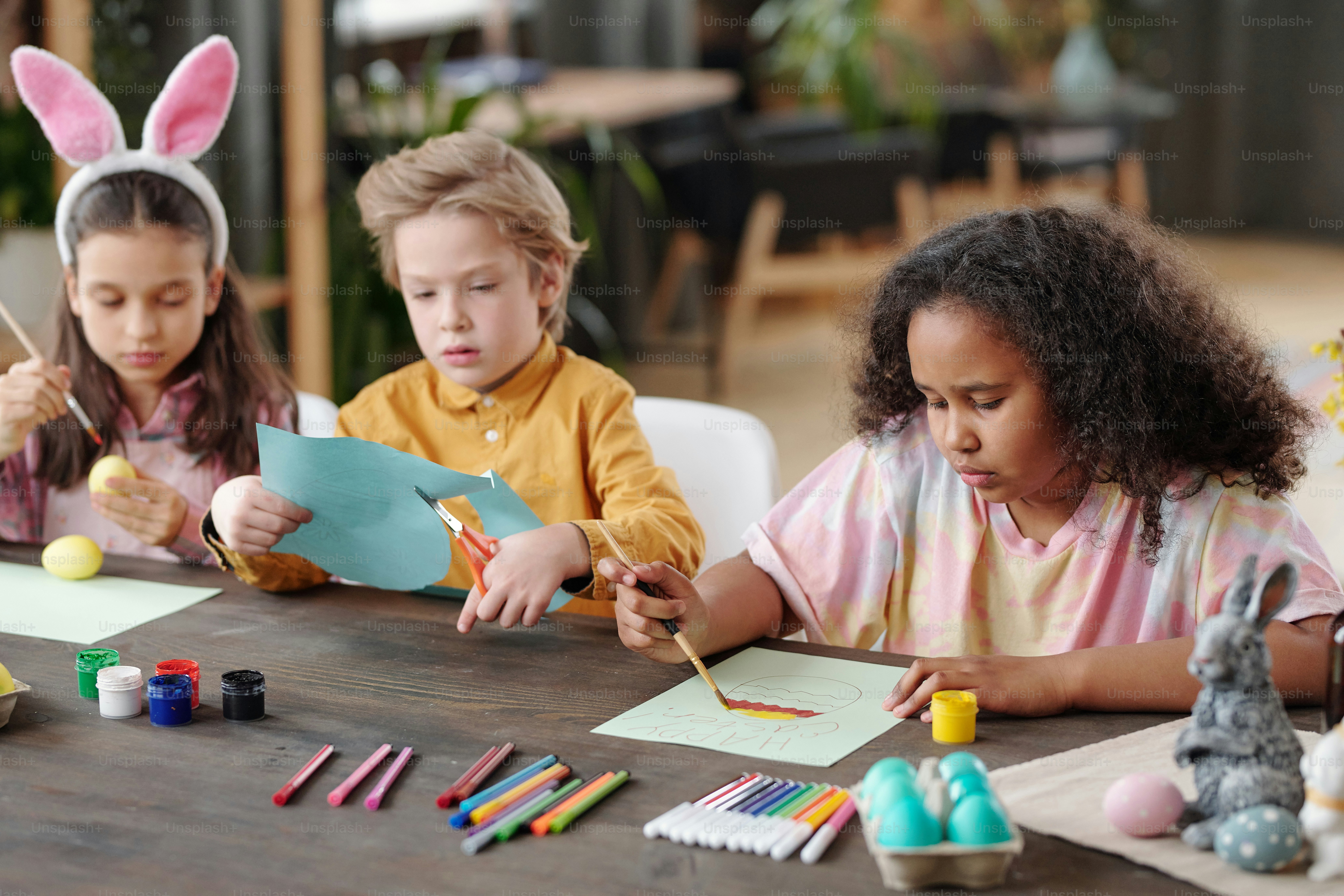 Group of three cute little friendly children of various ethnicities sitting by table in home environment and preparing presents for Easter