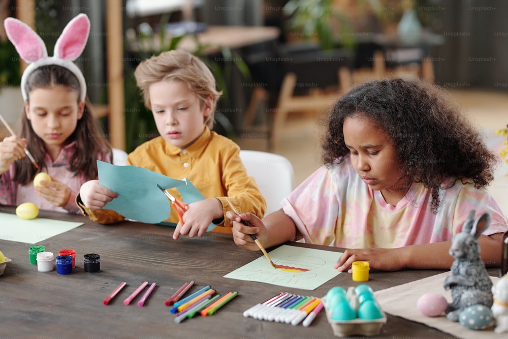 Group of three cute little friendly children of various ethnicities sitting by table in home environment and preparing presents for Easter