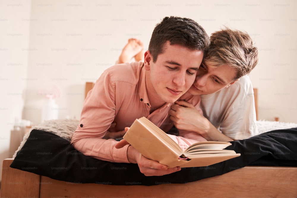 Was liest du? Zwei homosexuelle Menschen, die ein Buch auf dem Bett lesen. Glücklicher Mann und sein Freund umarmen sich beim Lesen. Konzept für schwule Beziehungen und Rechte. Stockfotografie