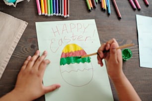 Hands of mixed-race little girl painting large Easter egg on blank paper among colorful highlighters and crayons while sitting by wooden table