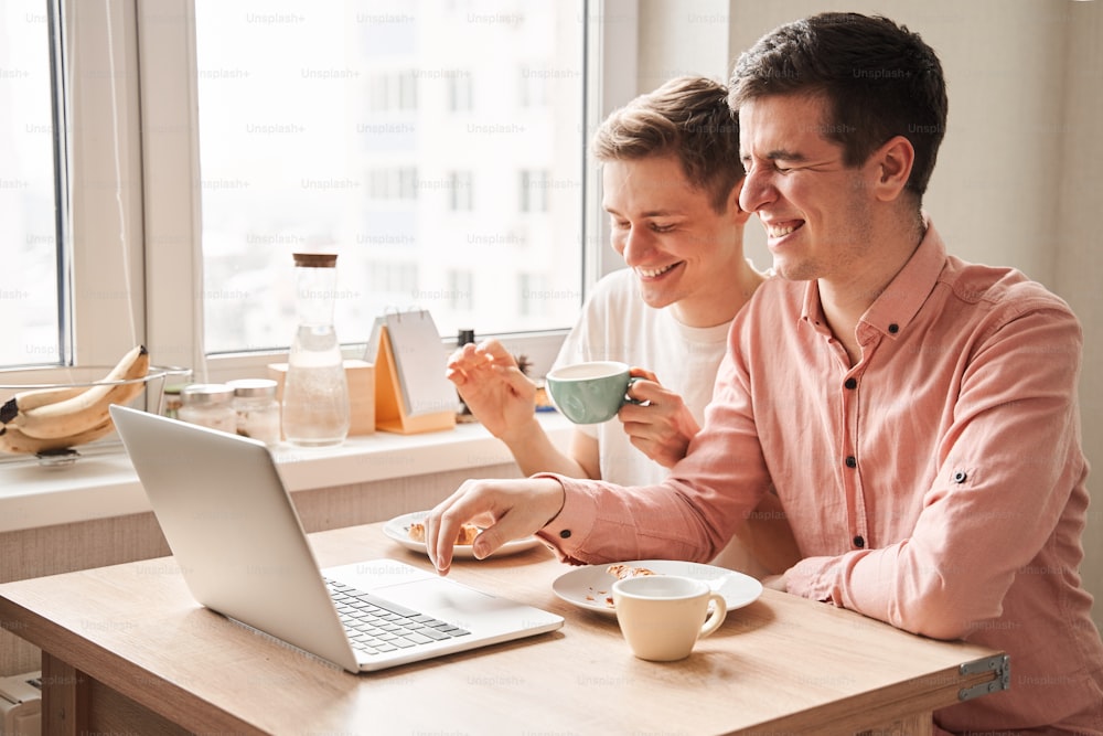 So funny. Waist up portrait view of the two gay man laughing out loud while watching something funny at the laptop while drinking morning coffee. Stock photo