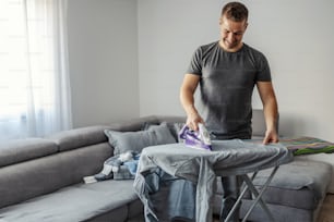 Home duty, ironing clothes. A man in his early thirties ironed clothes on an ironing board in the living room of a modern home during the day. Happy to contribute to household chores, happy to help