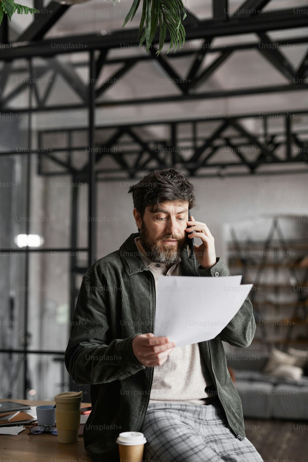 Vertical portrait of contemporary bearded businessman speaking by phone and holding documents in black office interior