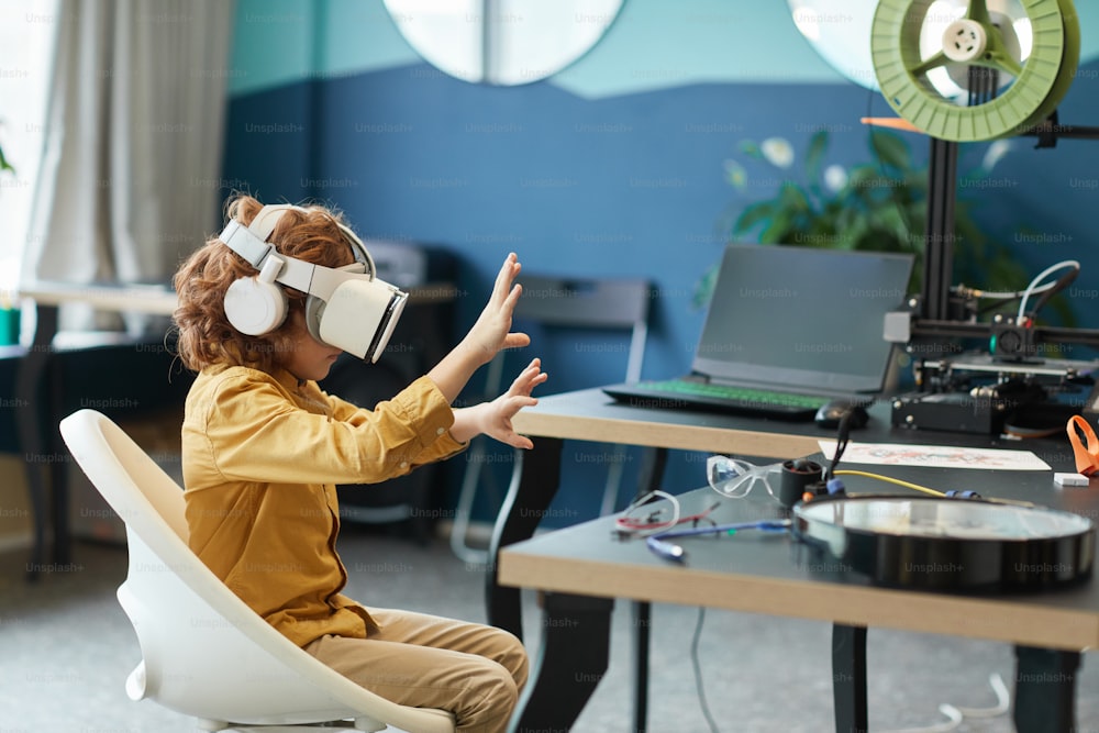 Seitenansichtsporträt eines kleinen Jungen, der ein VR-Headset trägt und die Hand ausstreckt, während er Augmented-Technologie im Schullabor testet, Kopierraum