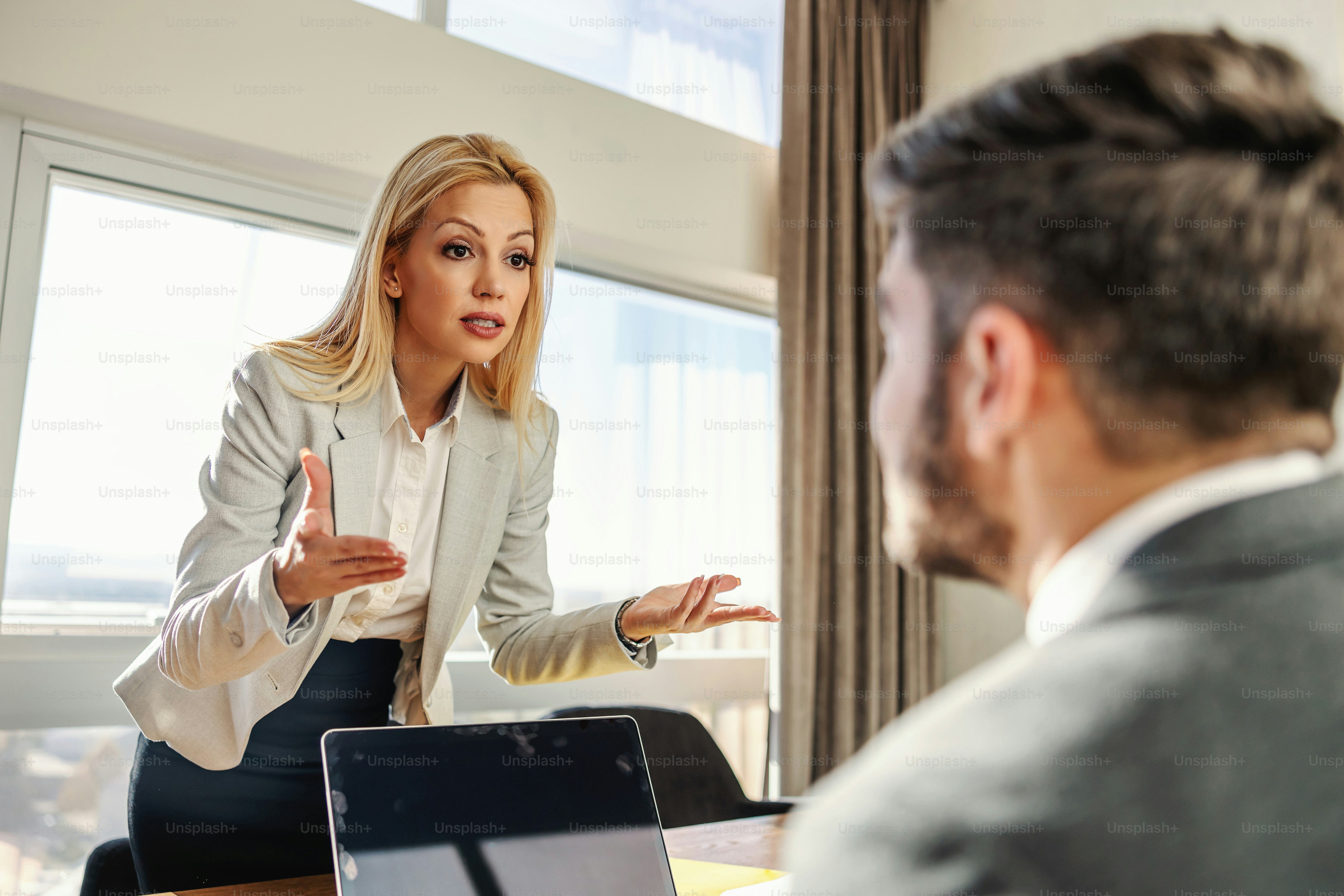 Blond adult businesswoman standing in the office with a laptop and having a fight with her colleague. Job must be done perfectly. Issues at work, argue