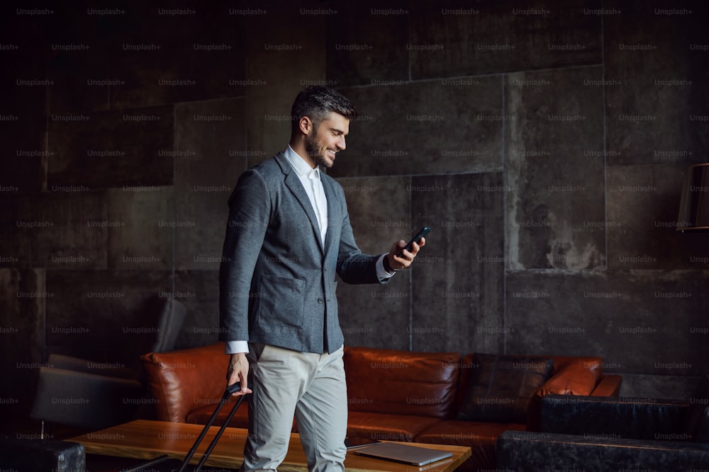 Happy businessman carrying his luggage while walking in the hall of a hotel and using his phone. Business trip, travel, technologies