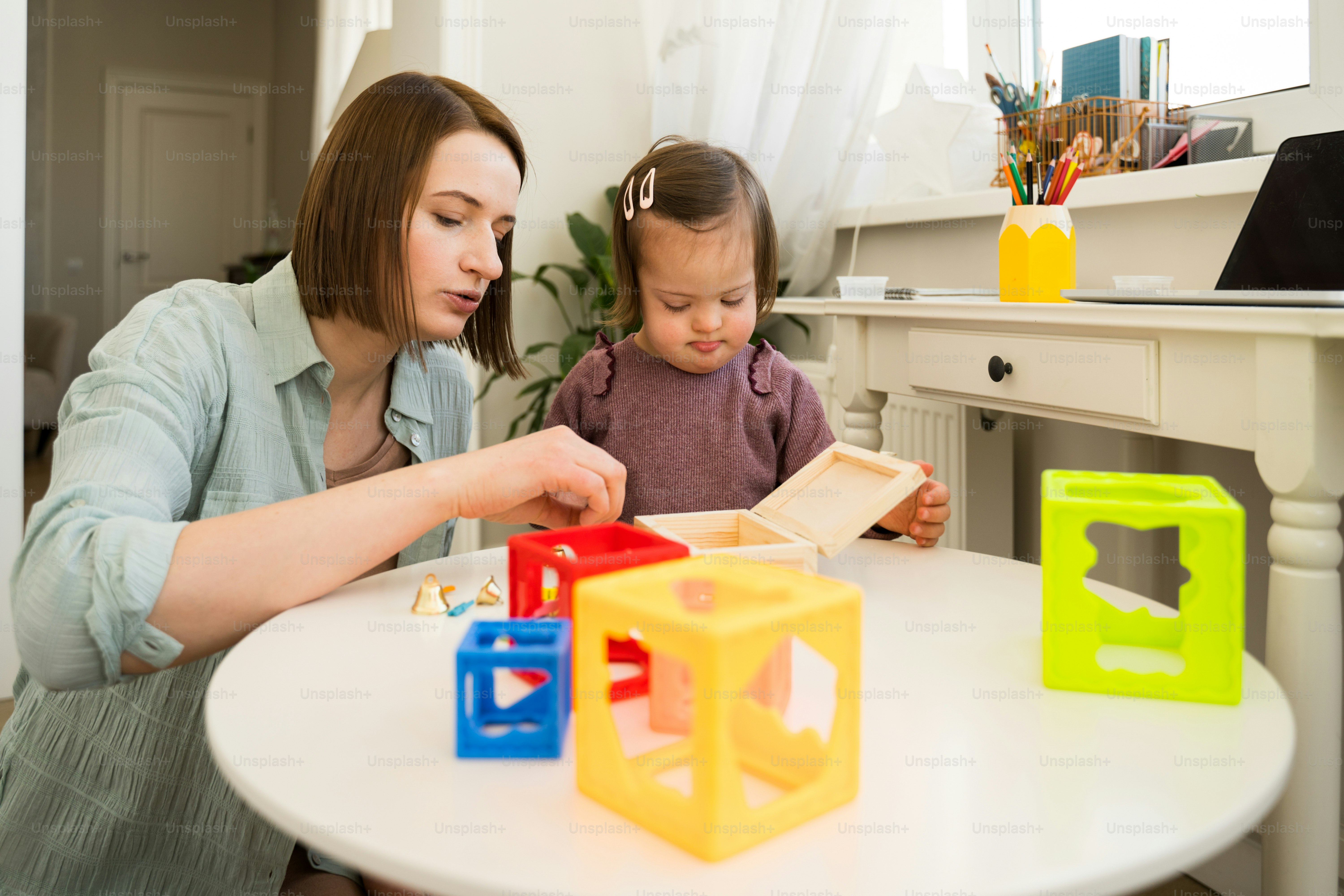 Porträtansicht des kleinen konzentrierten Mädchens mit Down-Syndrom, das mit seiner Mutter am Tisch mit ihrem Spielzeug spielt. Konzept für Menschen mit besonderen Bedürfnissen. Stockfotografie