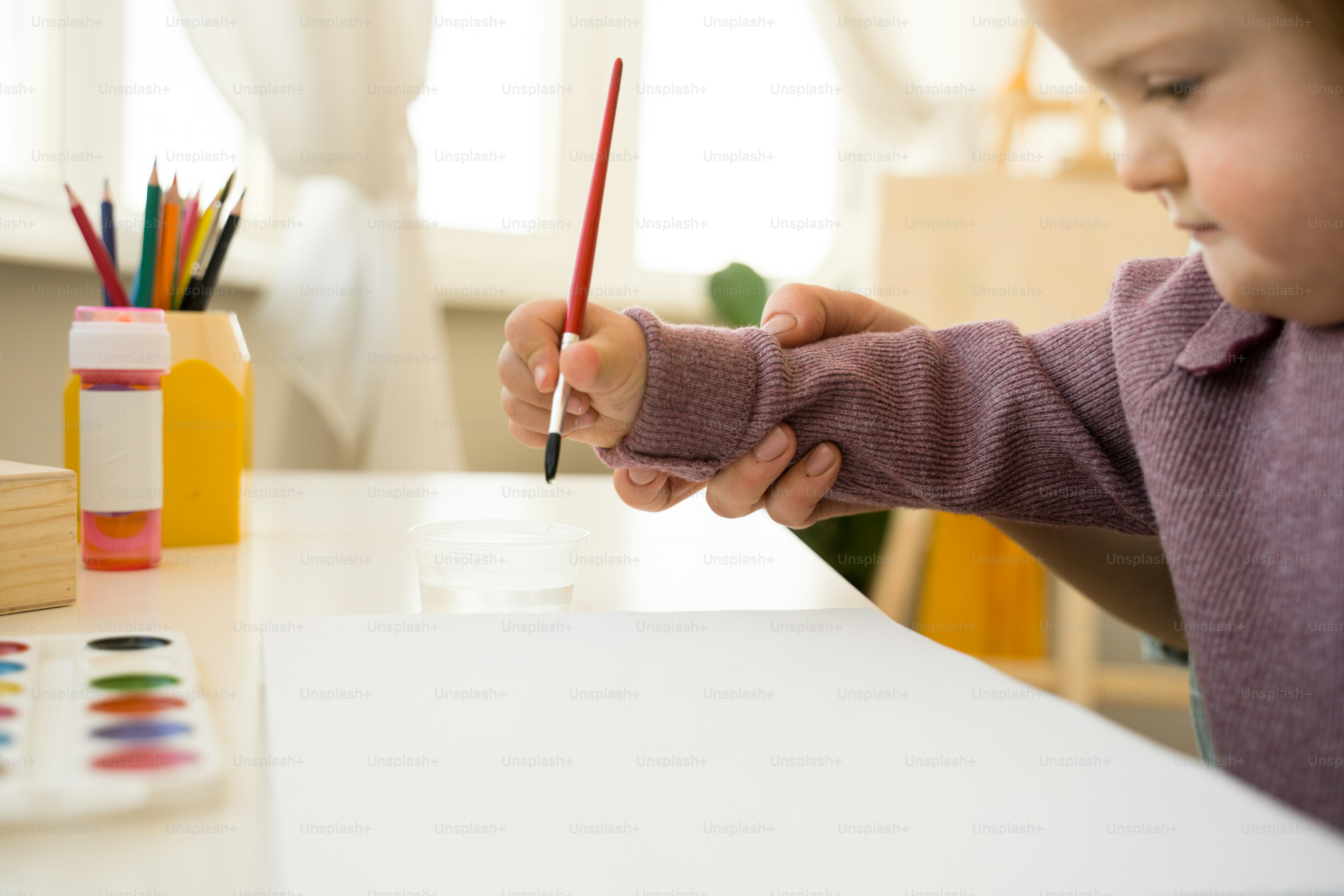 Nahaufnahme des weiblichen Kindes mit Down-Syndrom, das mit Farben auf Papier zeichnet, während es am Tisch an ihrem Arbeitsplatz sitzt. Mutter hilft beim Zeichnen ihrer Tochter. Stockfotografie
