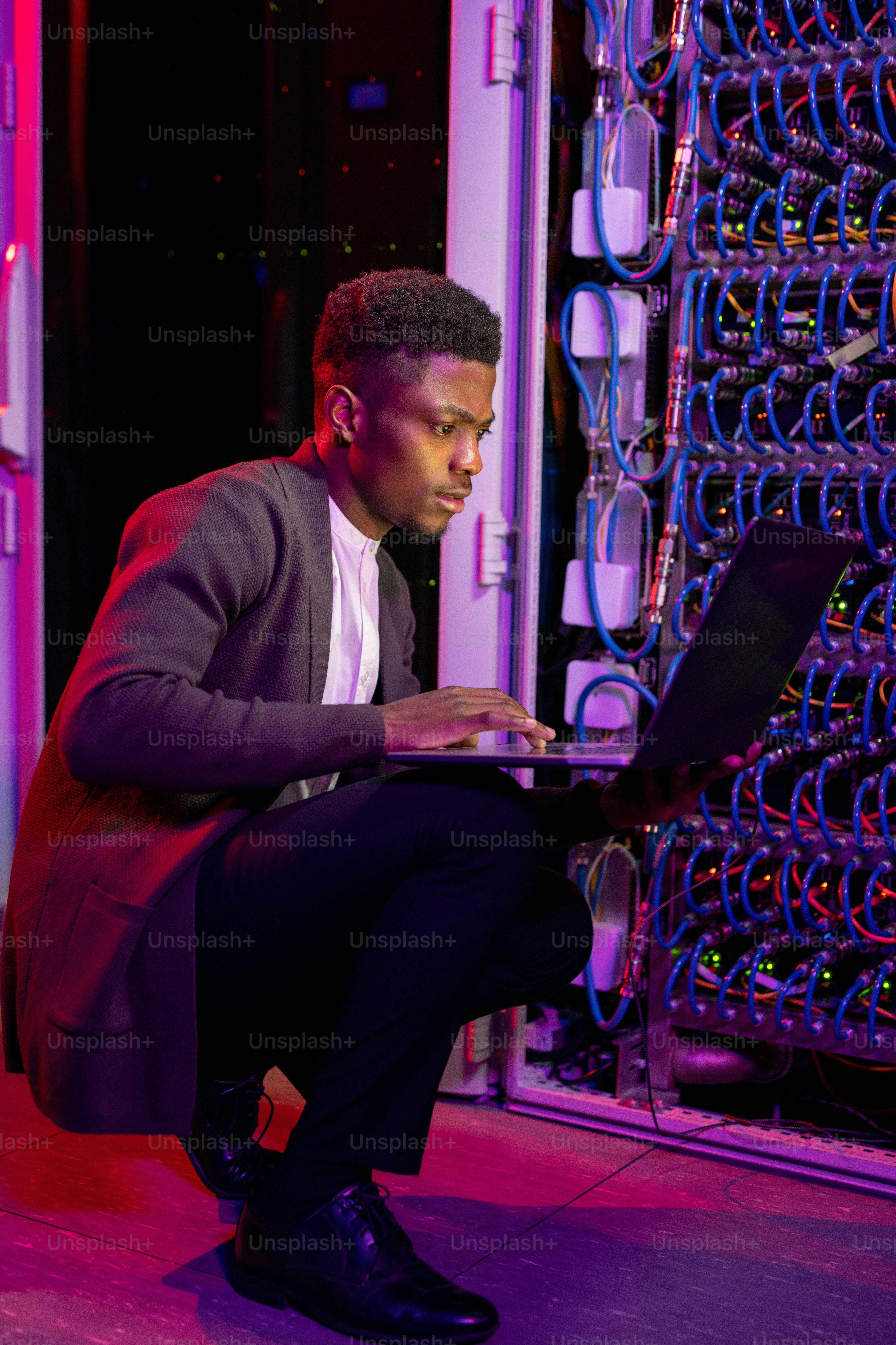Concentré jeune ingénieur informatique afro-américain accroupi près de l’armoire de serveur et analysant le travail du serveur à l’aide d’un ordinateur portable