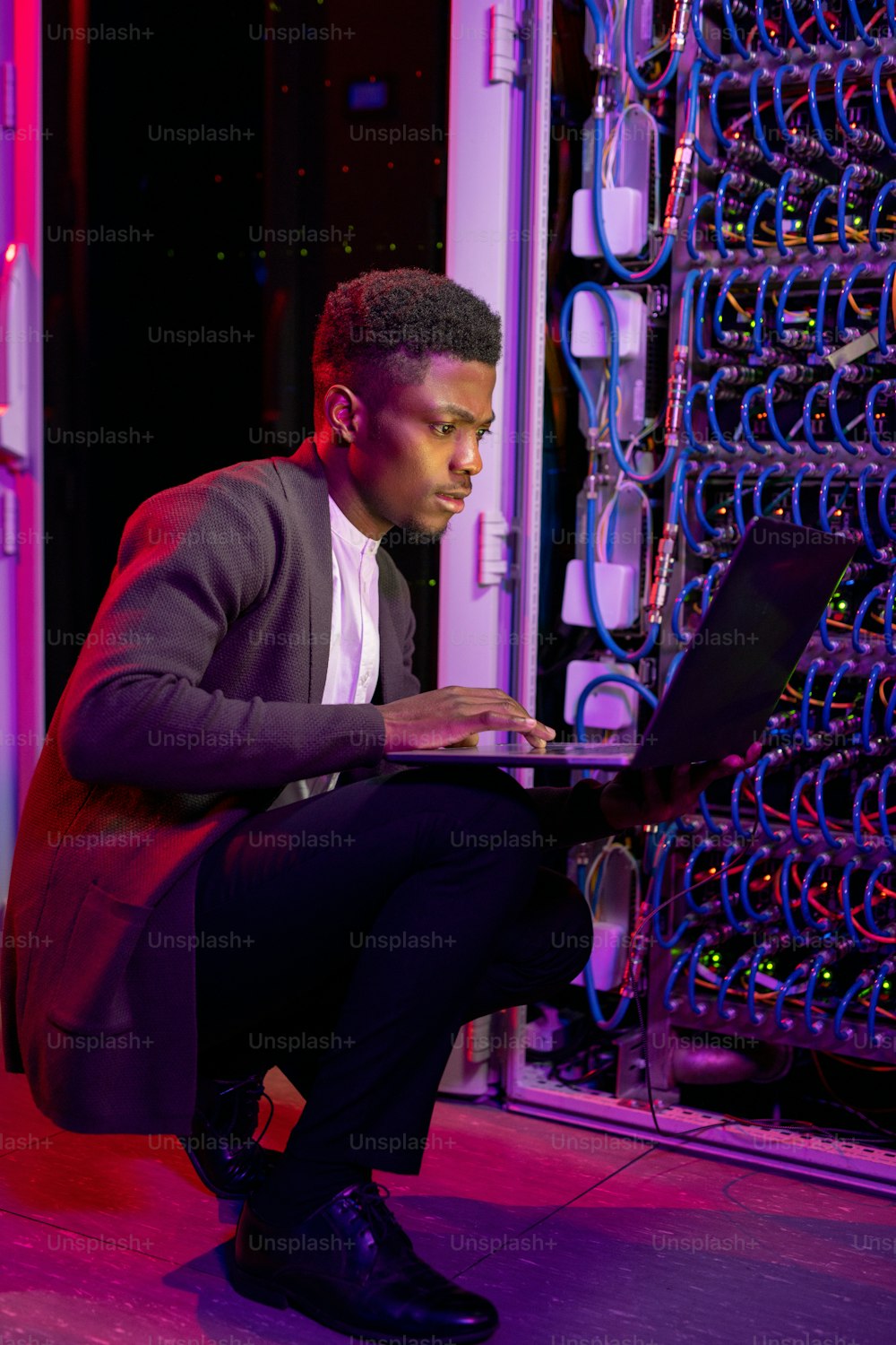 젊은 아프리카계 미국인 IT 엔지니어가 서버 캐비닛 옆에 웅크리고 앉아 노트북 컴퓨터를 사용하여 서버 작업을 분석하는 데 집중되어 있다