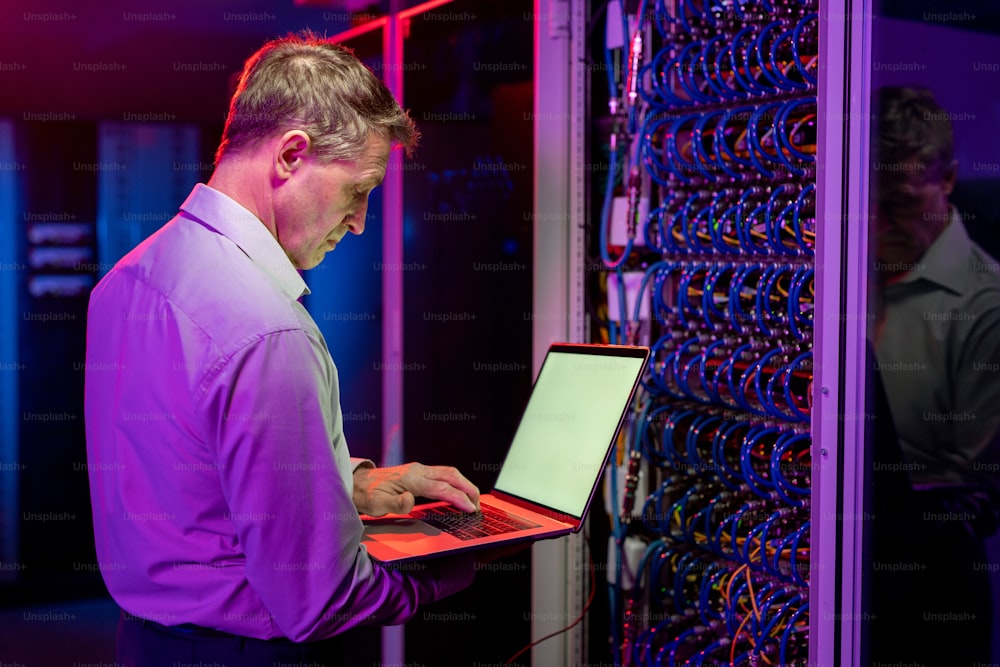 Ingegnere di rete serio e maturo in piedi all'armadio del server aperto e utilizzando il laptop durante la diagnosi dei problemi del server