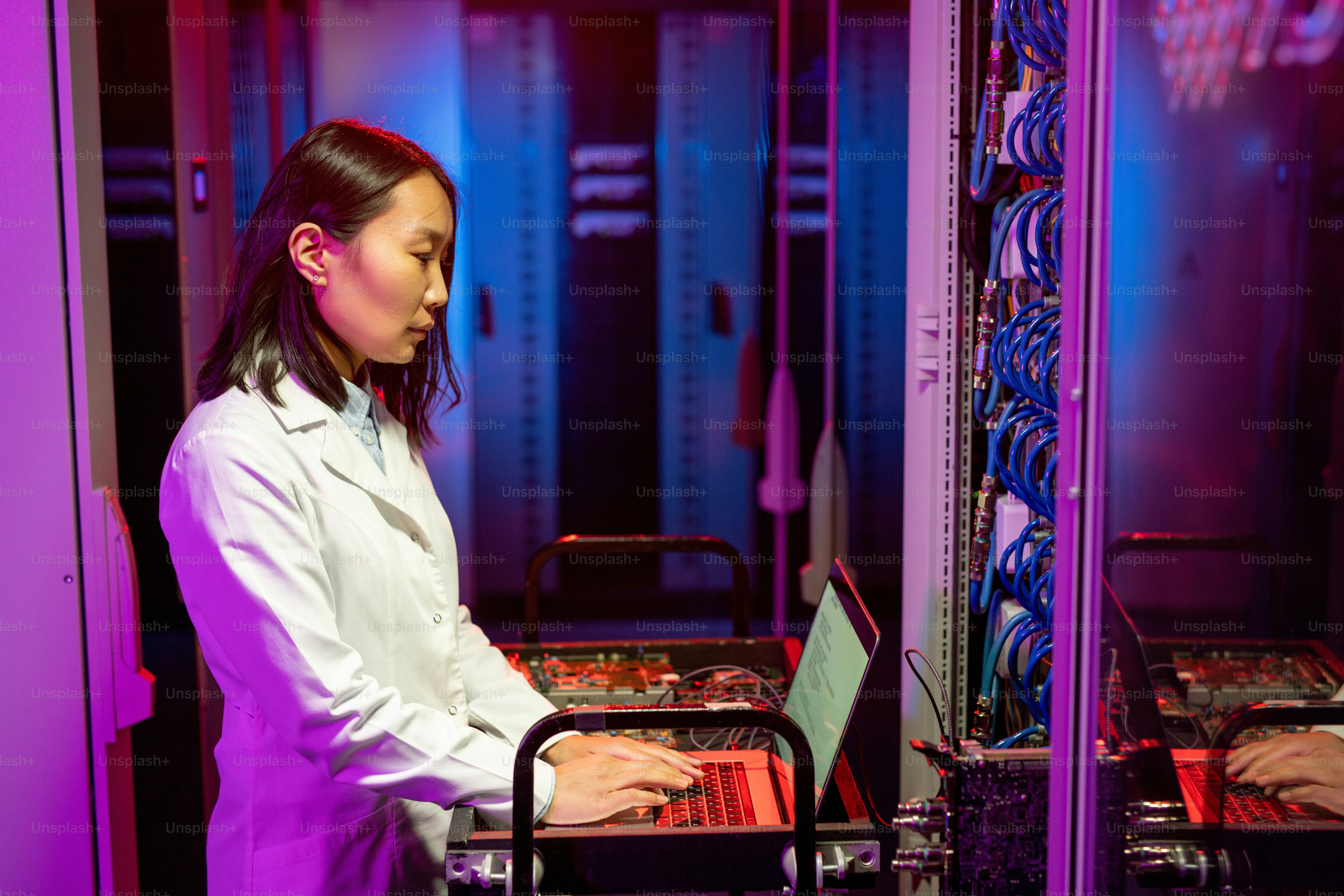 Serious young Asian IT specialist in white coat standing at cart and using laptop while performing diagnostic tests