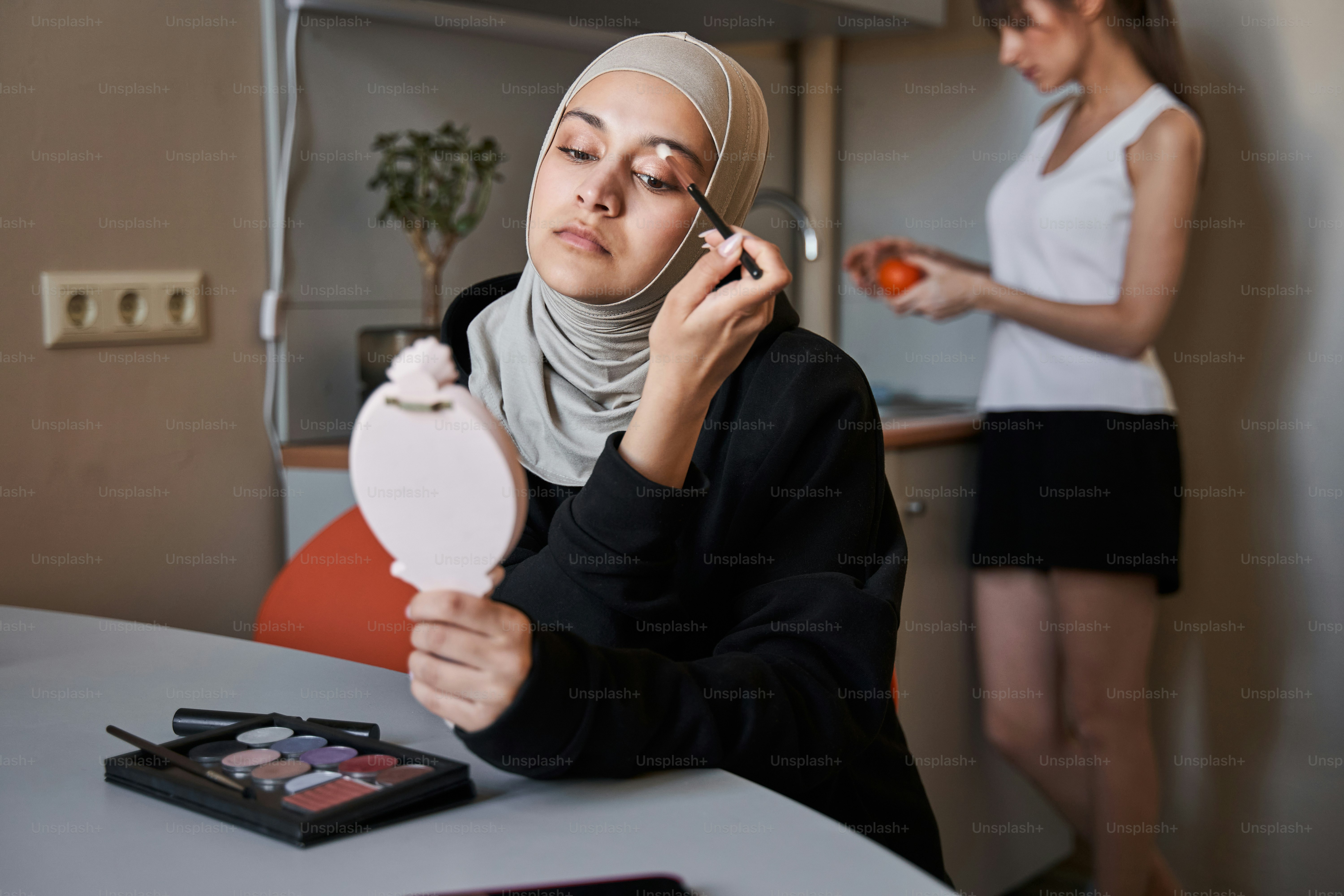 Portrait view of the gorgeous muslim woman looking at the hand mirror and applying shadows on her eyelids while her bestie washing fruits at the kitchen